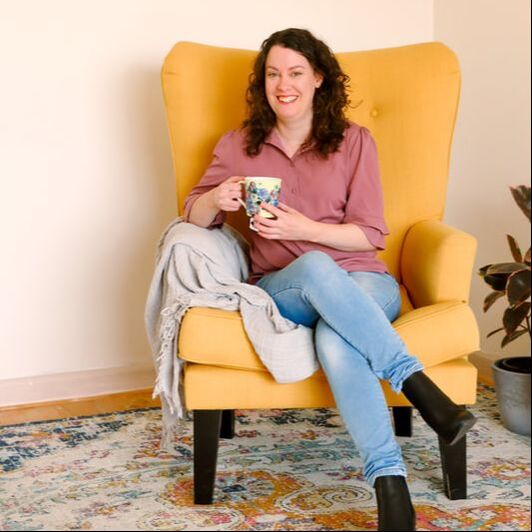 Online HAES Dietitian Melissa O'Loughlan is sitting on a yellow chair holding a cup of tea and smiling at the camera.